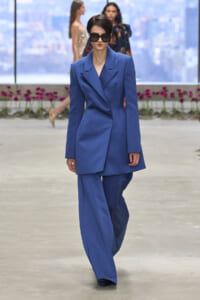 Model walks a runway in a royal blue tailored blazer and matching wide-leg pants, wearing oversized black sunglasses.