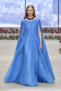 Model walks the runway in a light-blue satin gown with cape-like sleeves and a dark blue floral necklace, city backdrop behind.
