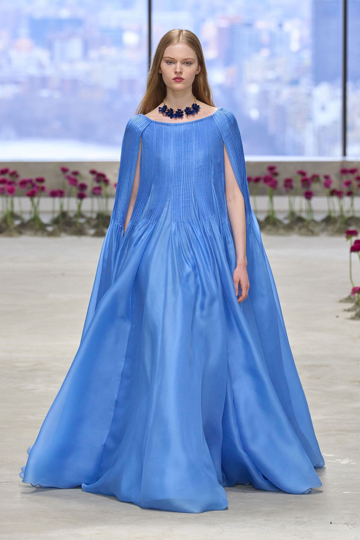 Model walks the runway in a light-blue satin gown with cape-like sleeves and a dark blue floral necklace, city backdrop behind.
