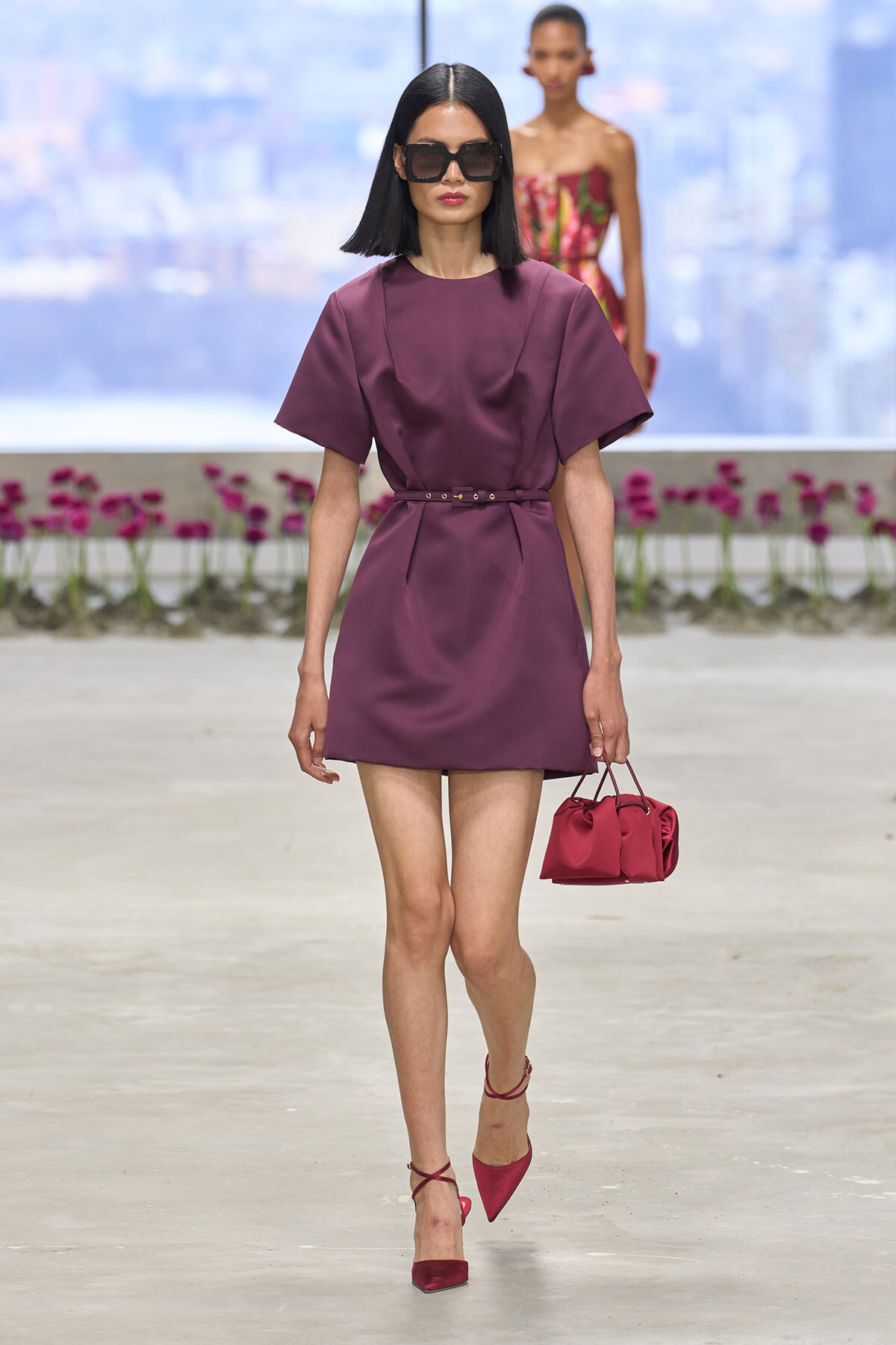 Model on a runway wearing a plum shift dress with a thin belt, red heels, and a red handbag, accessorized with oversized sunglasses.