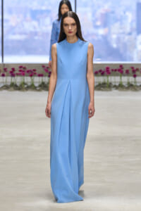 Model walks the runway in a light blue sleeveless gown with front pleats and a long flowing skirt.