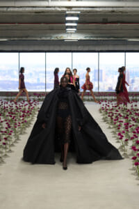 Model in a dramatic black cape gown walks down a fashion-show runway lined with pink flowers, with other models and a city skyline visible through large windows in the background.