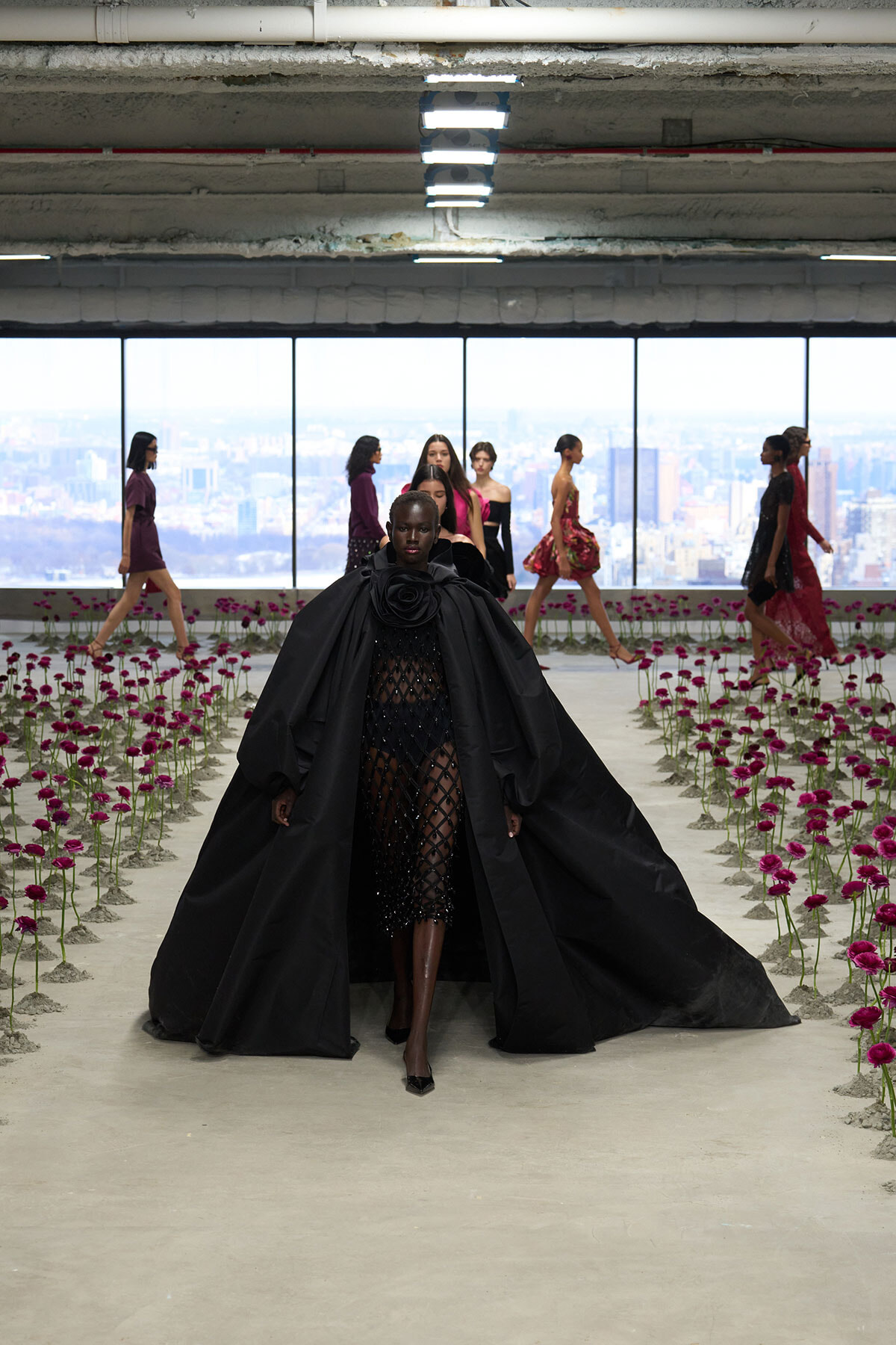 Model in a dramatic black cape gown walks down a fashion-show runway lined with pink flowers, with other models and a city skyline visible through large windows in the background.