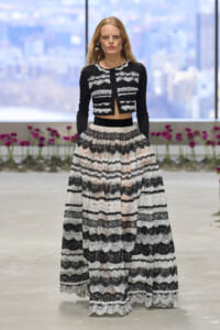 Model walking a fashion show wearing a black cropped lace jacket and a full-length black-and-white striped lace skirt.