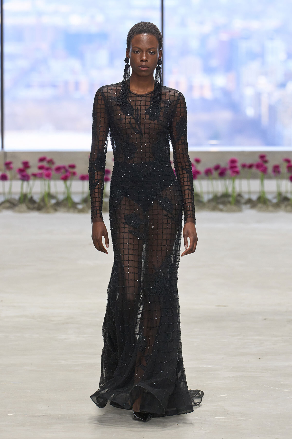 Model walks a runway in a floor-length sheer black beaded gown with a grid pattern and long sleeves.