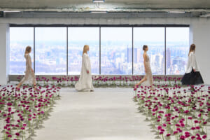 Models walk a minimalist runway indoors with a city skyline visible through large windows, flanked by rows of magenta flowers.