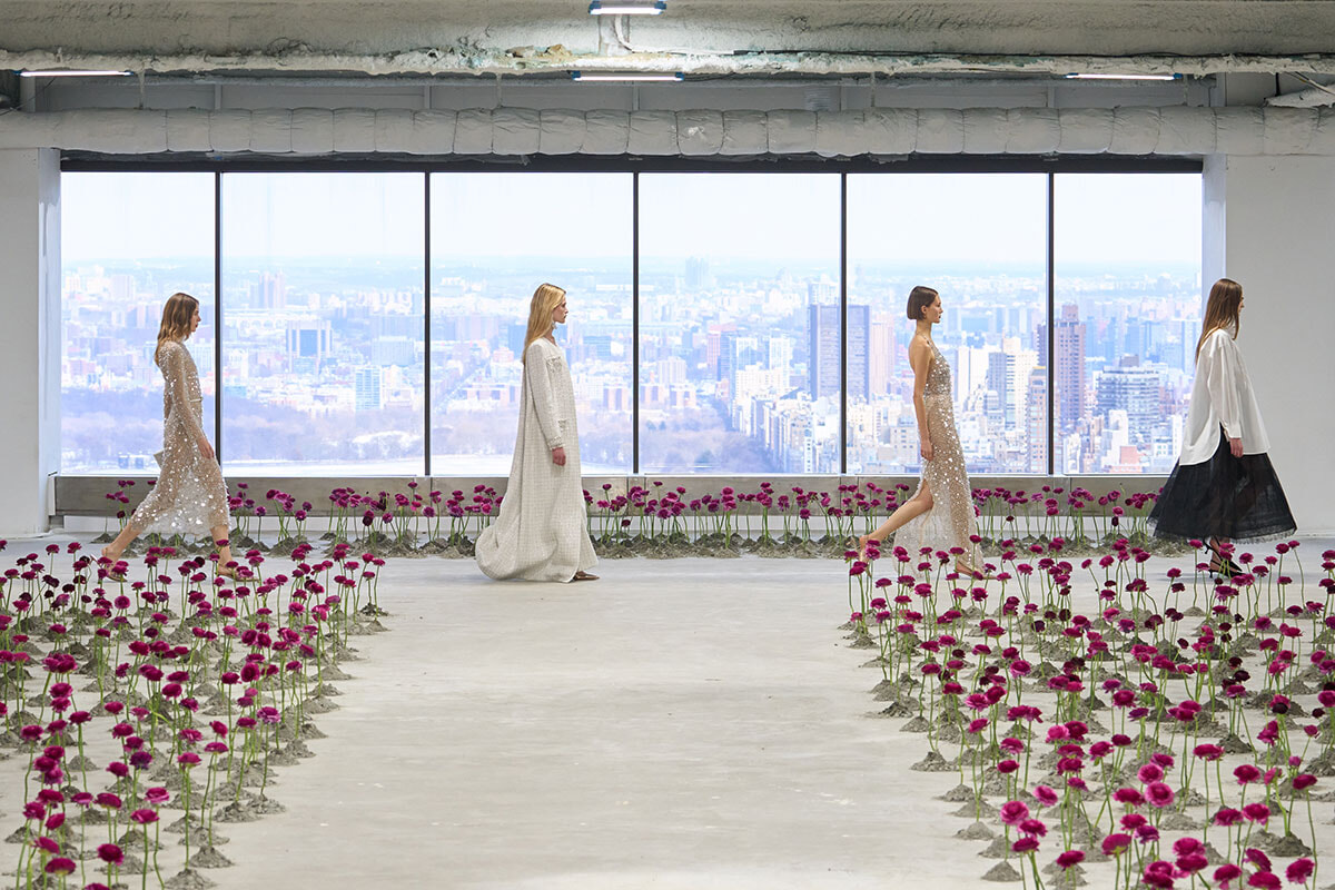 Models walk a minimalist runway indoors with a city skyline visible through large windows, flanked by rows of magenta flowers.