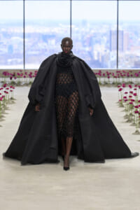 Model walking the runway in a dramatic black cape with voluminous sleeves and a sheer beaded dress, city skyline in the background.