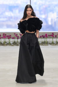 Runway model in a dramatic black off-shoulder top with oversized rose-embellished sleeves and wide black pants.