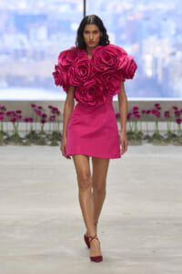 Model wearing a vivid pink mini dress with oversized rose-shaped appliqués at the shoulders, walking on a runway.