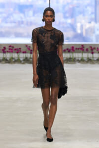 Runway model in a sheer black grid dress with floral appliqués, walking a fashion show runway.