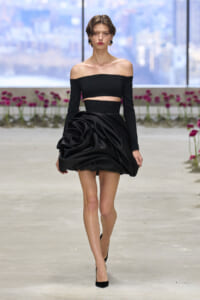 Model walking a fashion show in a black off-shoulder crop top and dramatic satin draped mini skirt, paired with black heels.
