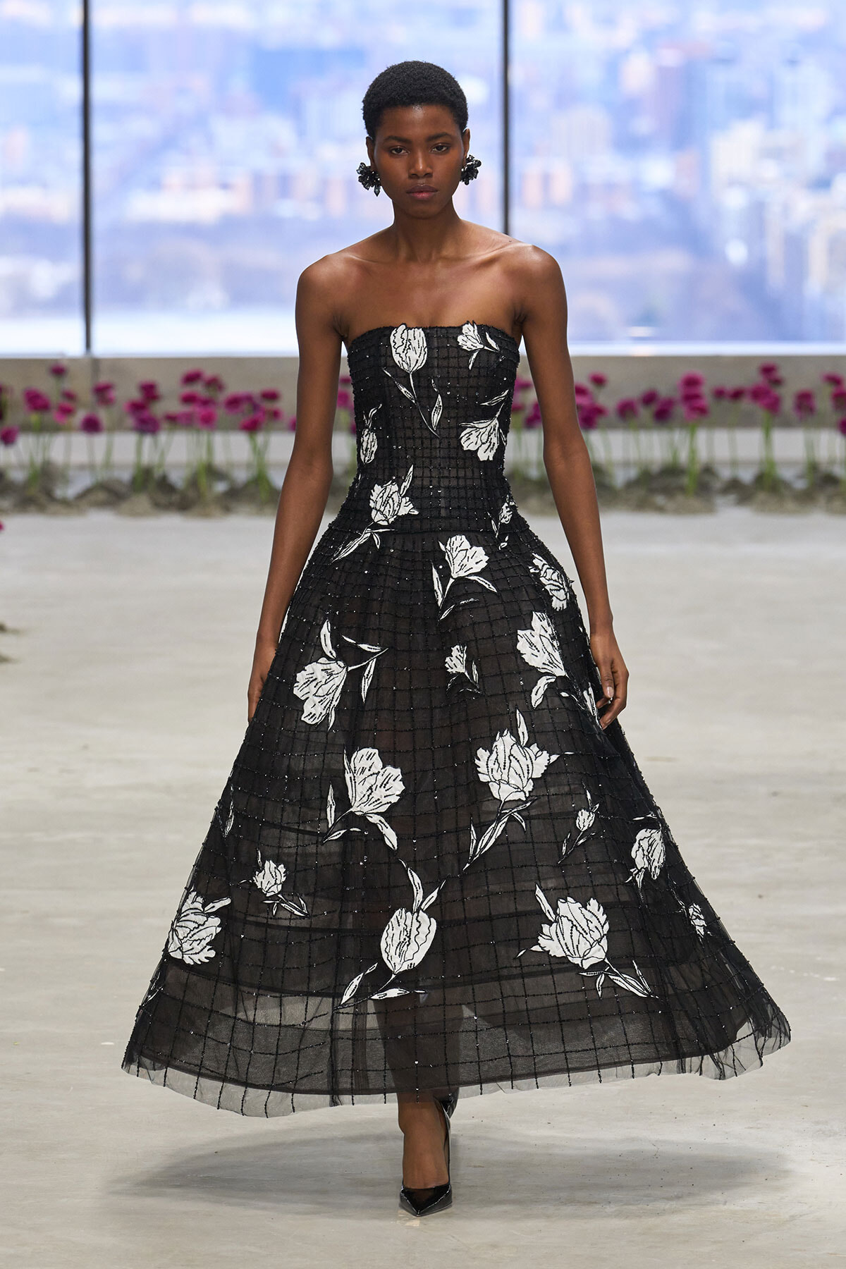 Strapless black gown with white floral print and grid overlay, model walking on runway indoors with city backdrop