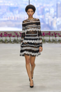 Model walking on an indoor runway in a black-and-white lace long-sleeve dress with sheer panels and scalloped stripes.