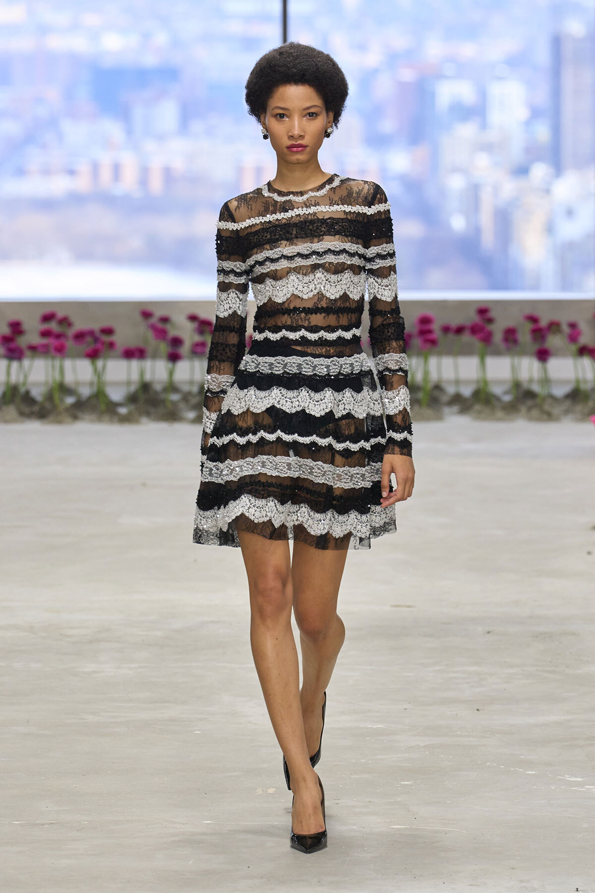 Model walking on an indoor runway in a black-and-white lace long-sleeve dress with sheer panels and scalloped stripes.
