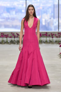 Model walks runway in a bright pink sleeveless gown with a deep V neckline and voluminous mermaid skirt, black heels visible at the hem.