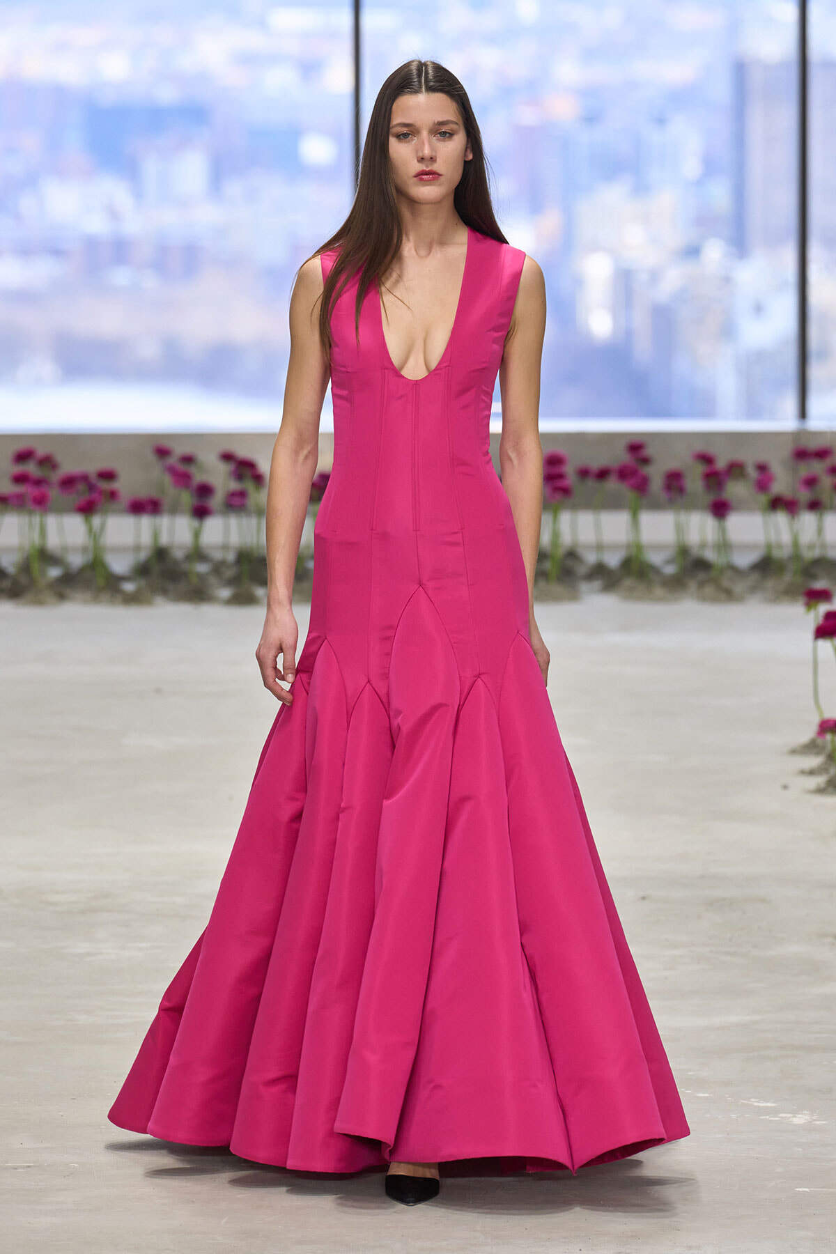 Model walks runway in a bright pink sleeveless gown with a deep V neckline and voluminous mermaid skirt, black heels visible at the hem.