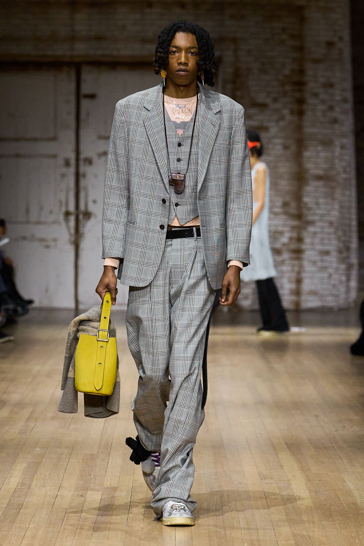 Male model in an oversized gray plaid suit with vest, walking a runway while holding a bright yellow bag and draped coat.
