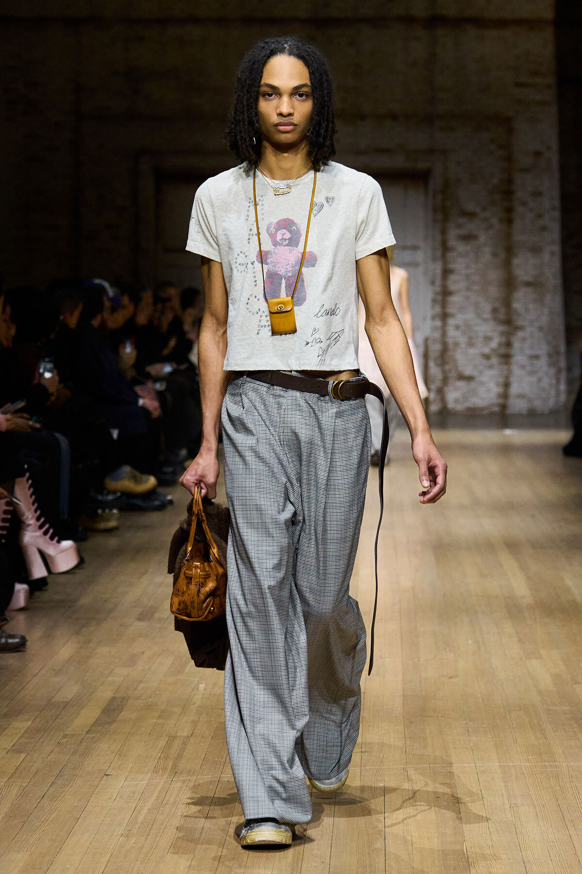 Model walks a fashion runway wearing a light gray graphic T-shirt tucked into wide, gray plaid trousers with a brown belt, carrying a small brown handbag and wearing a long necklace; audience visible in background.