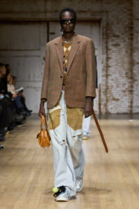 Model walks a runway in a brown plaid blazer with patchwork jeans and an orange tan handbag, wearing dark sunglasses.