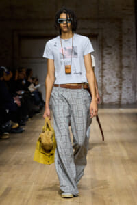 Model walks a runway in a light blue graphic T-shirt, wide plaid gray trousers, brown belt, and oversized blue sunglasses; carries a yellow fuzzy jacket and a small brown pouch.