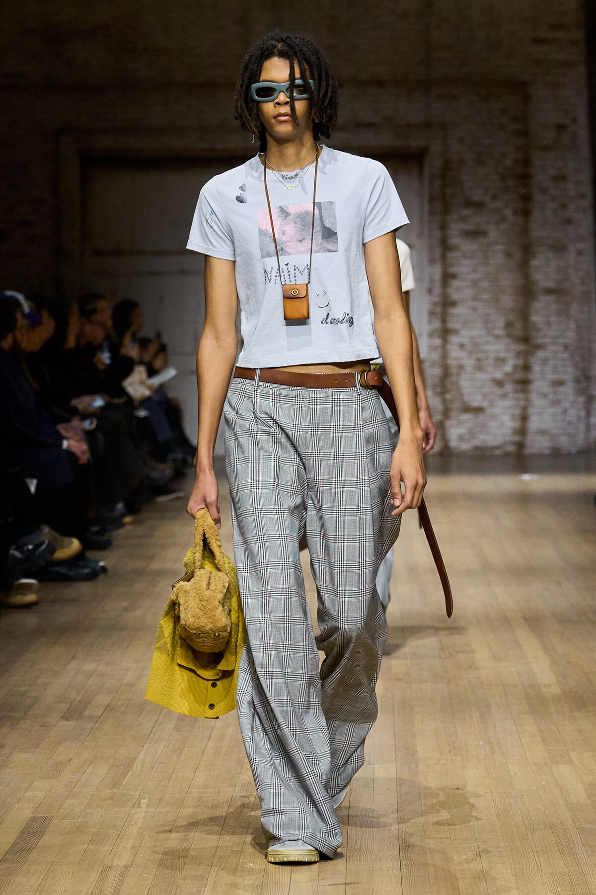 Model walks a runway in a light blue graphic T-shirt, wide plaid gray trousers, brown belt, and oversized blue sunglasses; carries a yellow fuzzy jacket and a small brown pouch.