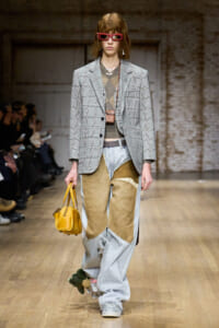 Model on a runway wearing a gray plaid blazer over an oversized patchwork jeans look, with pink sunglasses and a yellow handbag.