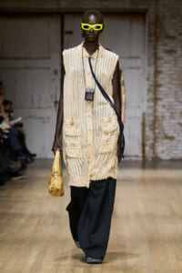 Model on a runway wearing a cream striped sleeveless coat with large pockets, paired with wide-leg dark pants and neon yellow sunglasses. She carries a small brown crossbody bag and a yellow doodled handbag.