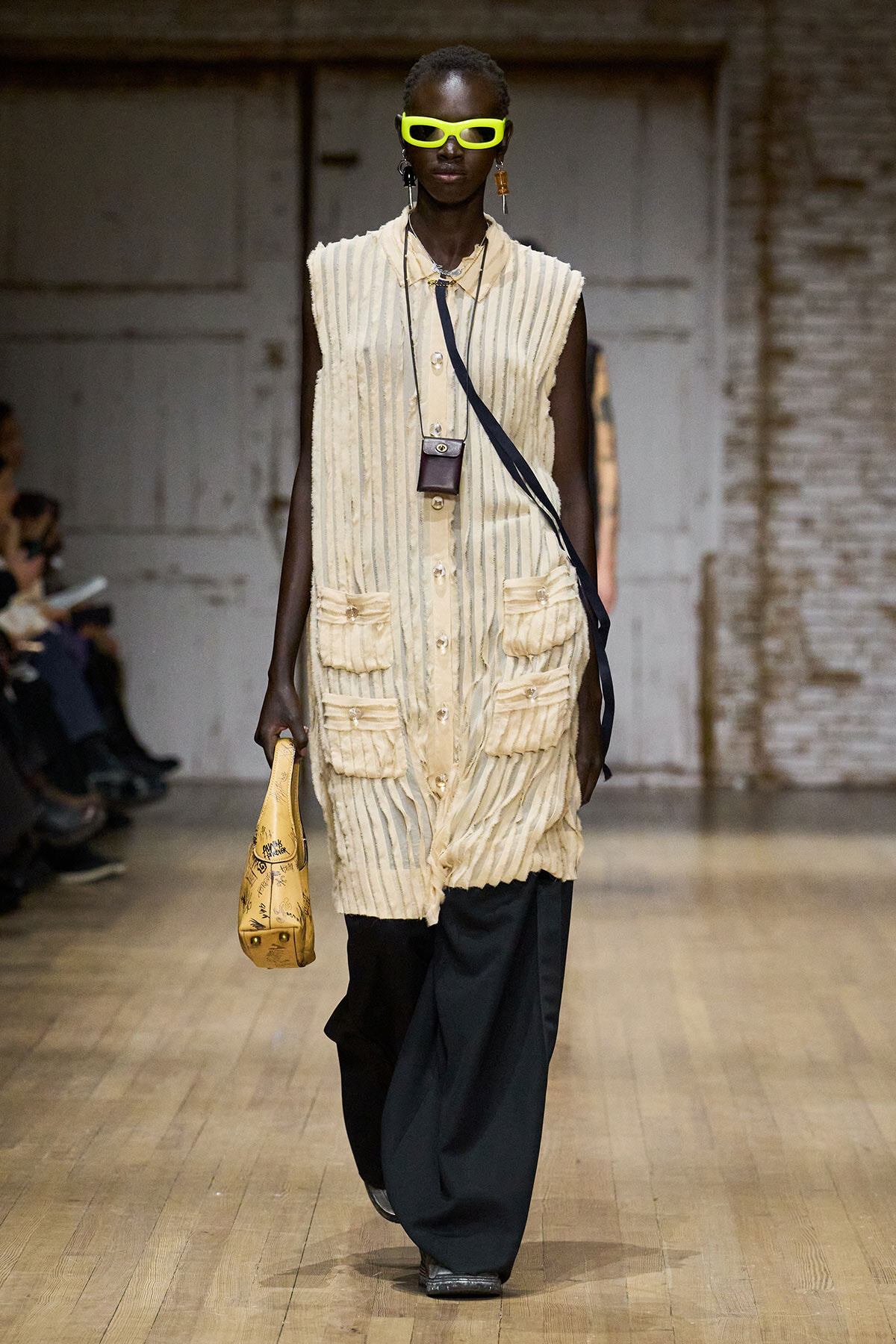 Model on a runway wearing a cream striped sleeveless coat with large pockets, paired with wide-leg dark pants and neon yellow sunglasses. She carries a small brown crossbody bag and a yellow doodled handbag.