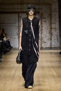 Model walking a fashion runway in a sheer black sleeveless coat with large pockets over wide-leg black pants, wearing futuristic sunglasses and a small crossbody bag.