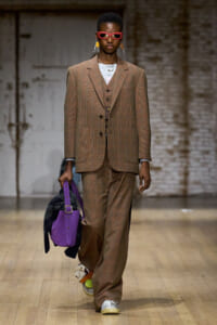 Model wearing a brown plaid three-piece suit with red sunglasses, carrying a purple handbag on a wooden runway.
