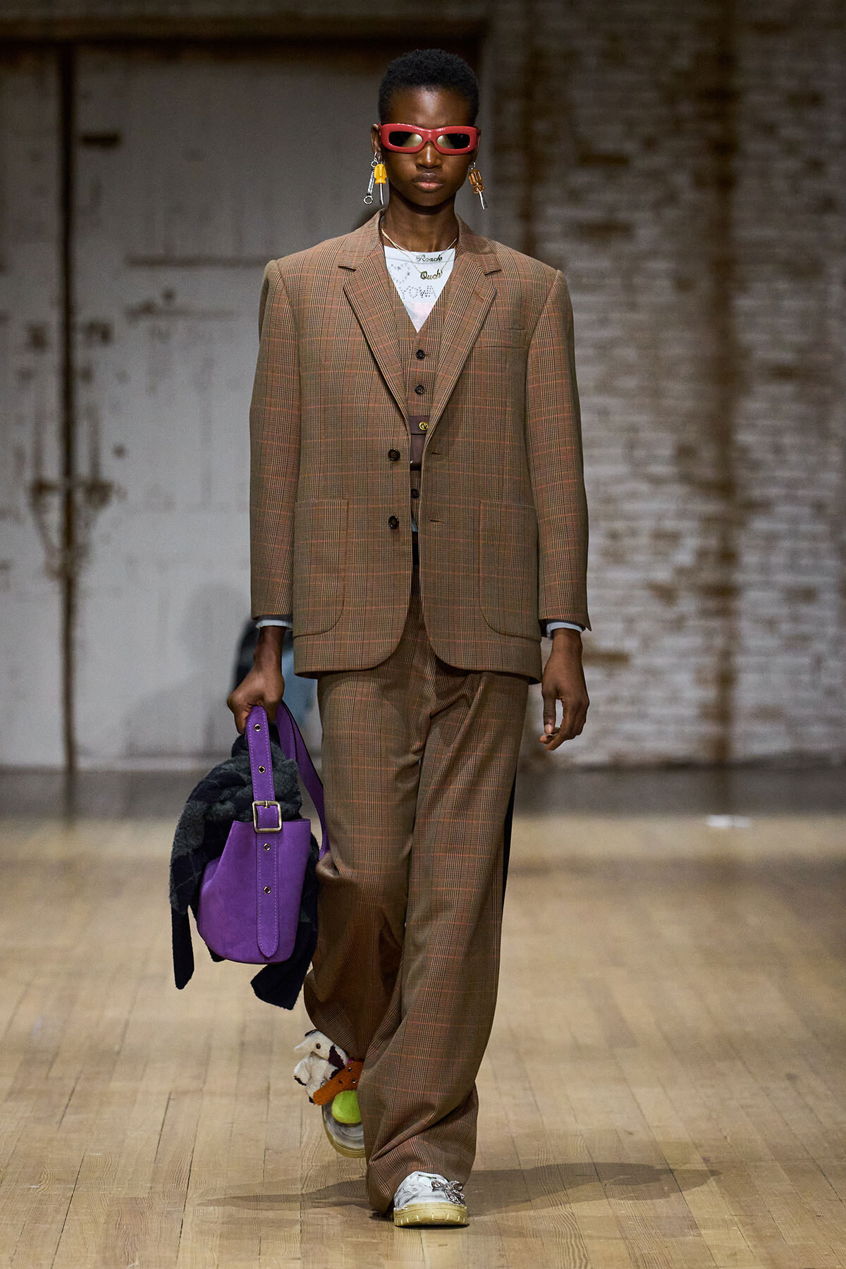 Model wearing a brown plaid three-piece suit with red sunglasses, carrying a purple handbag on a wooden runway.