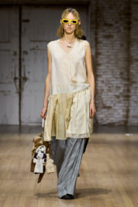 Runway model in a cream sleeveless blouse with a draped tie and layered ivory skirt over gray plaid trousers, wearing yellow sunglasses and carrying a plush toy bag.