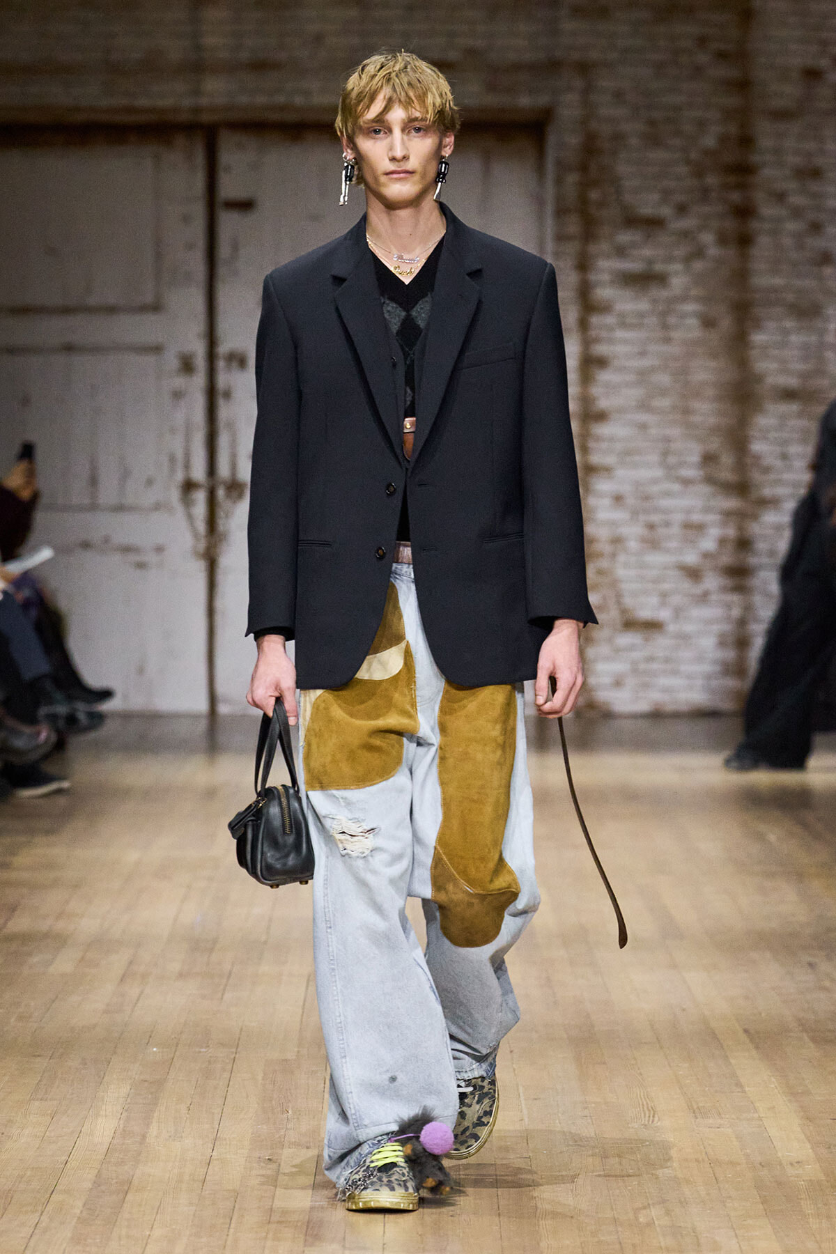 Male model on a runway wearing an oversized black blazer, patchwork jeans with mustard patches, leopard sneakers with a pink pom-pom, and a small black handbag.