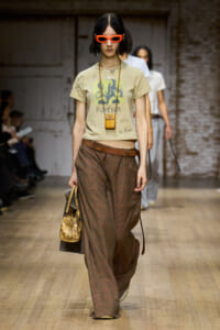 Runway model in a beige graphic tee with 'FOREVER', brown plaid wide-leg pants, brown belt, orange sunglasses, and a small tan crossbody bag.