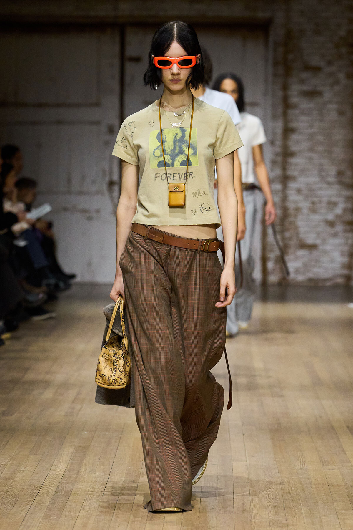 Runway model in a beige graphic tee with 'FOREVER', brown plaid wide-leg pants, brown belt, orange sunglasses, and a small tan crossbody bag.