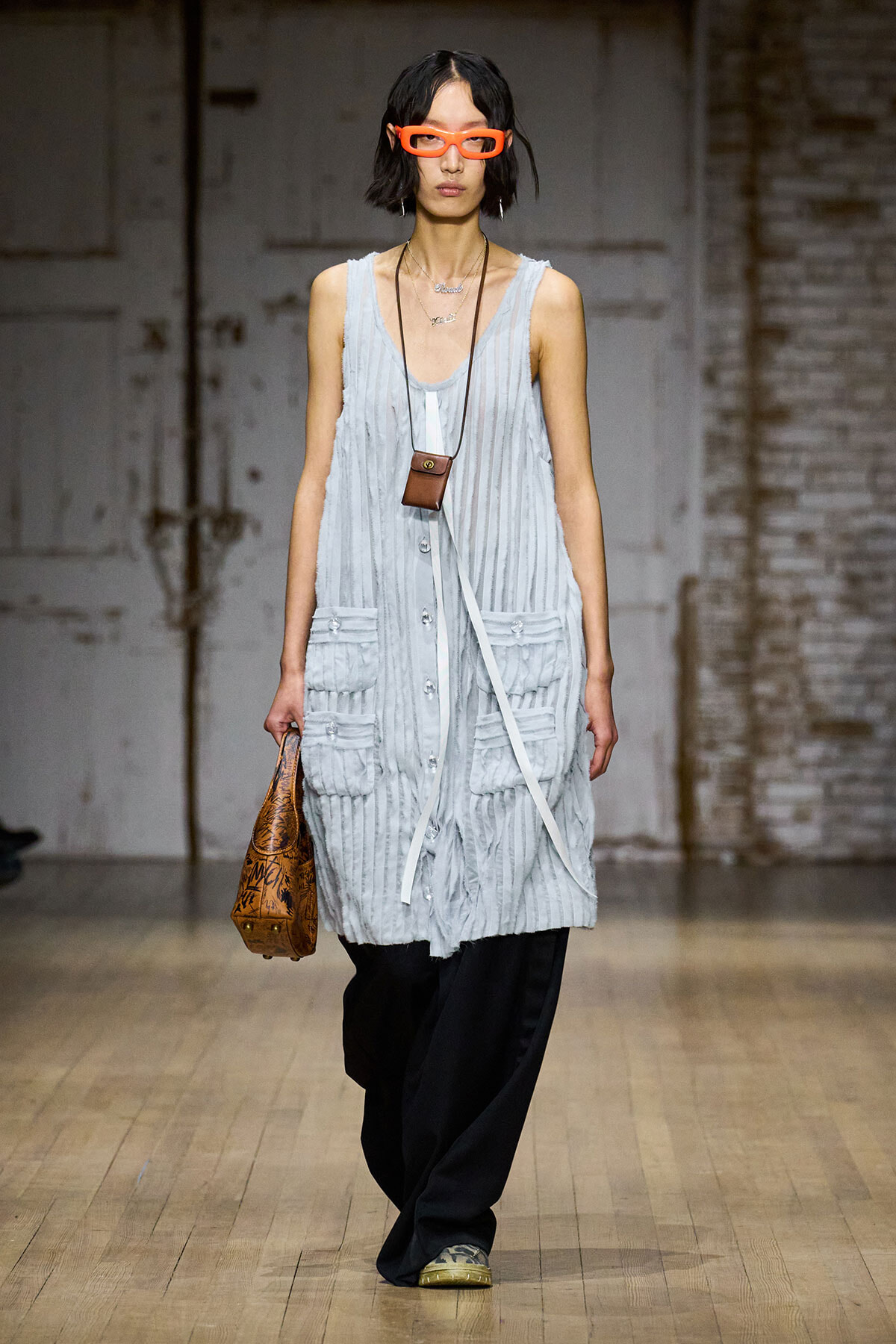 Model on runway wearing a light gray sleeveless tunic with pockets, black wide-leg pants, bright orange glasses, and layered necklaces, carrying a small brown crossbody bag and a patterned brown handbag.