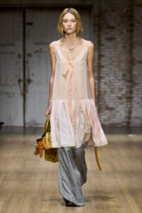 Runway model in a pale pink sheer dress with a tied ribbon over plaid gray trousers, carrying a tan bag with a plush carrot charm.
