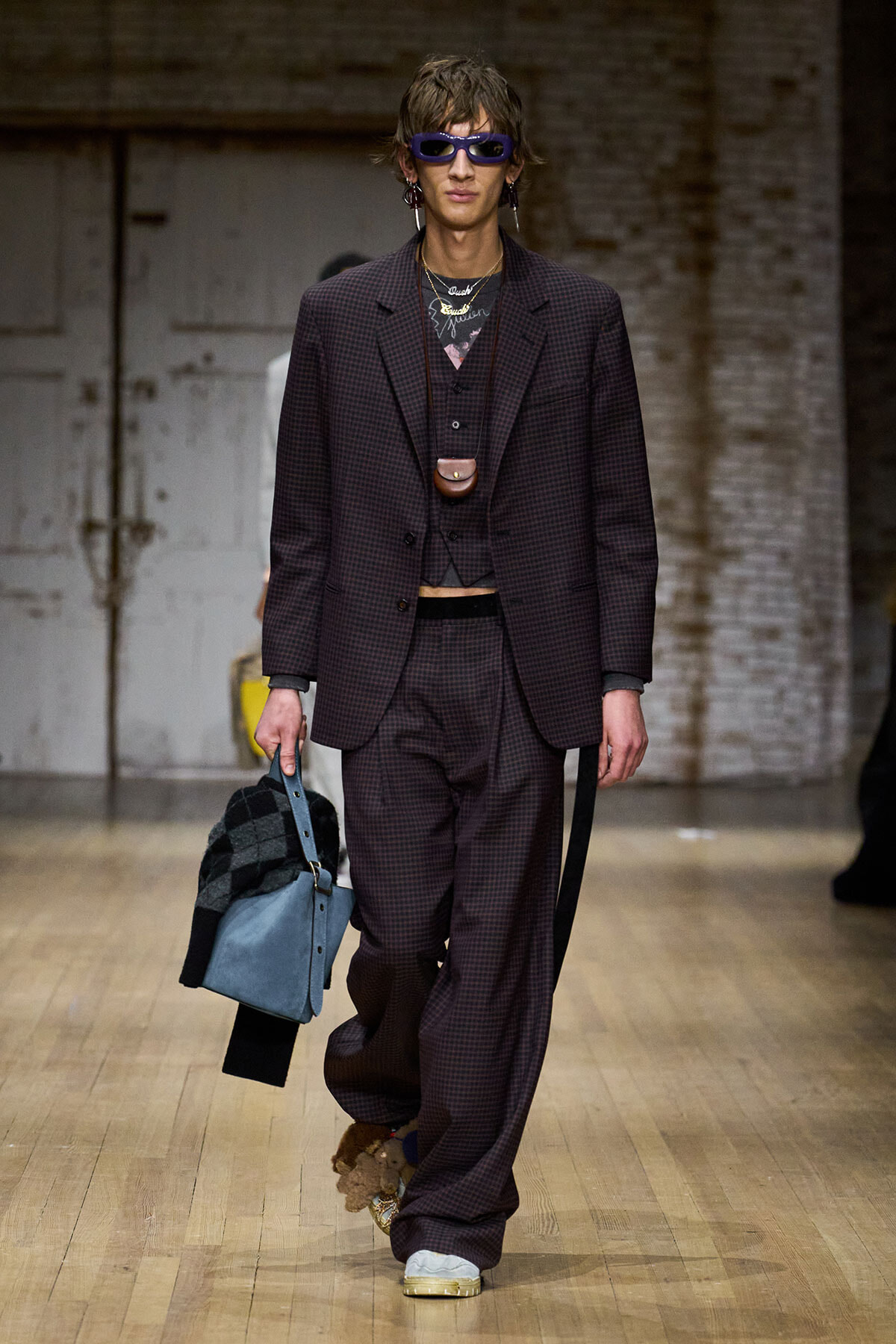 Male model in a burgundy checkered suit with oversized blazer, vest, and baggy trousers, wearing purple sunglasses and a small pendant pouch at the chest, walking a runway with a blue tote bag.",
