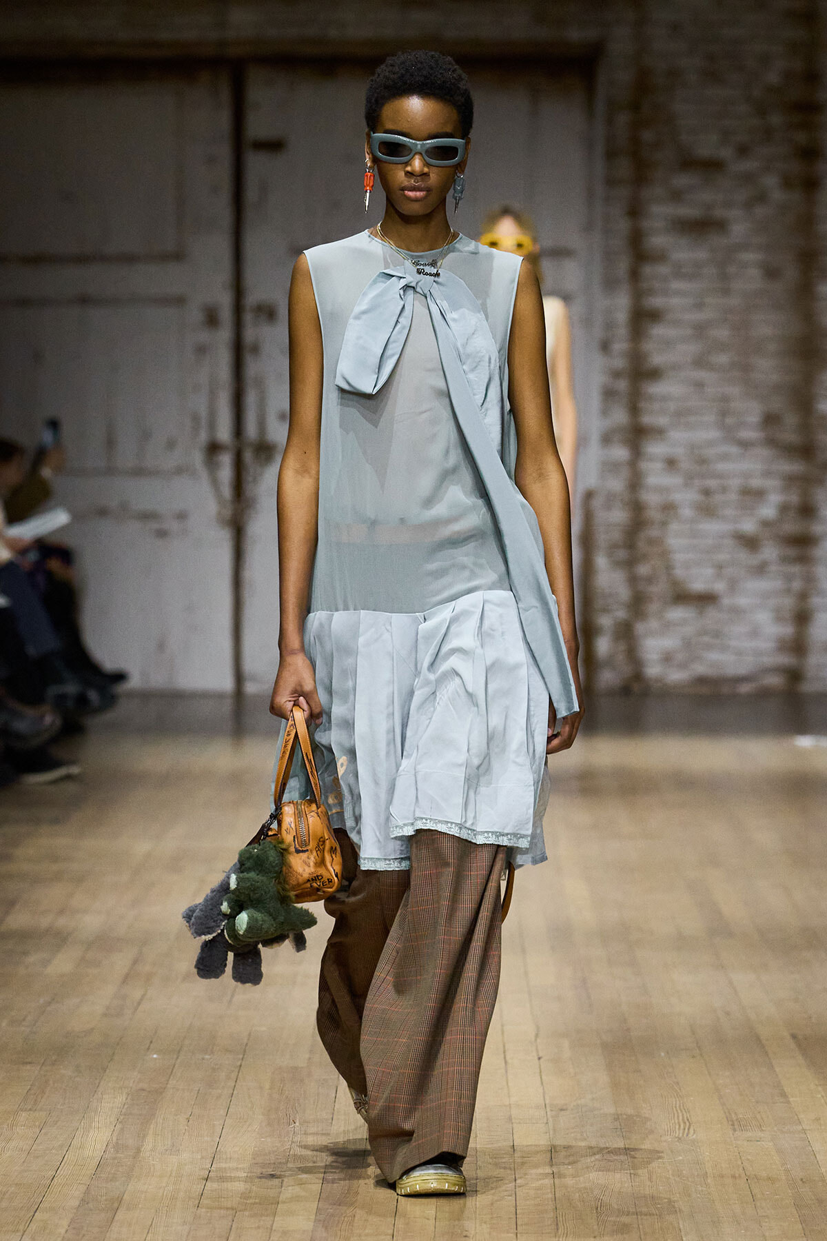 Model walking a fashion runway in a light blue sleeveless tunic with bow detail, paired with wide brown plaid trousers and a small tan handbag with plush keychain accessories