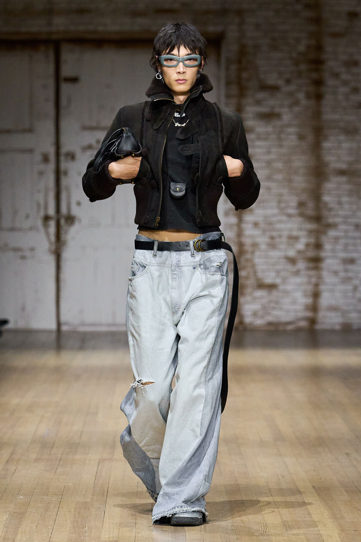 Model walking a runway in a black cropped jacket, oversized light-wash jeans, blue sunglasses, and chunky accessories, carrying a black bag.