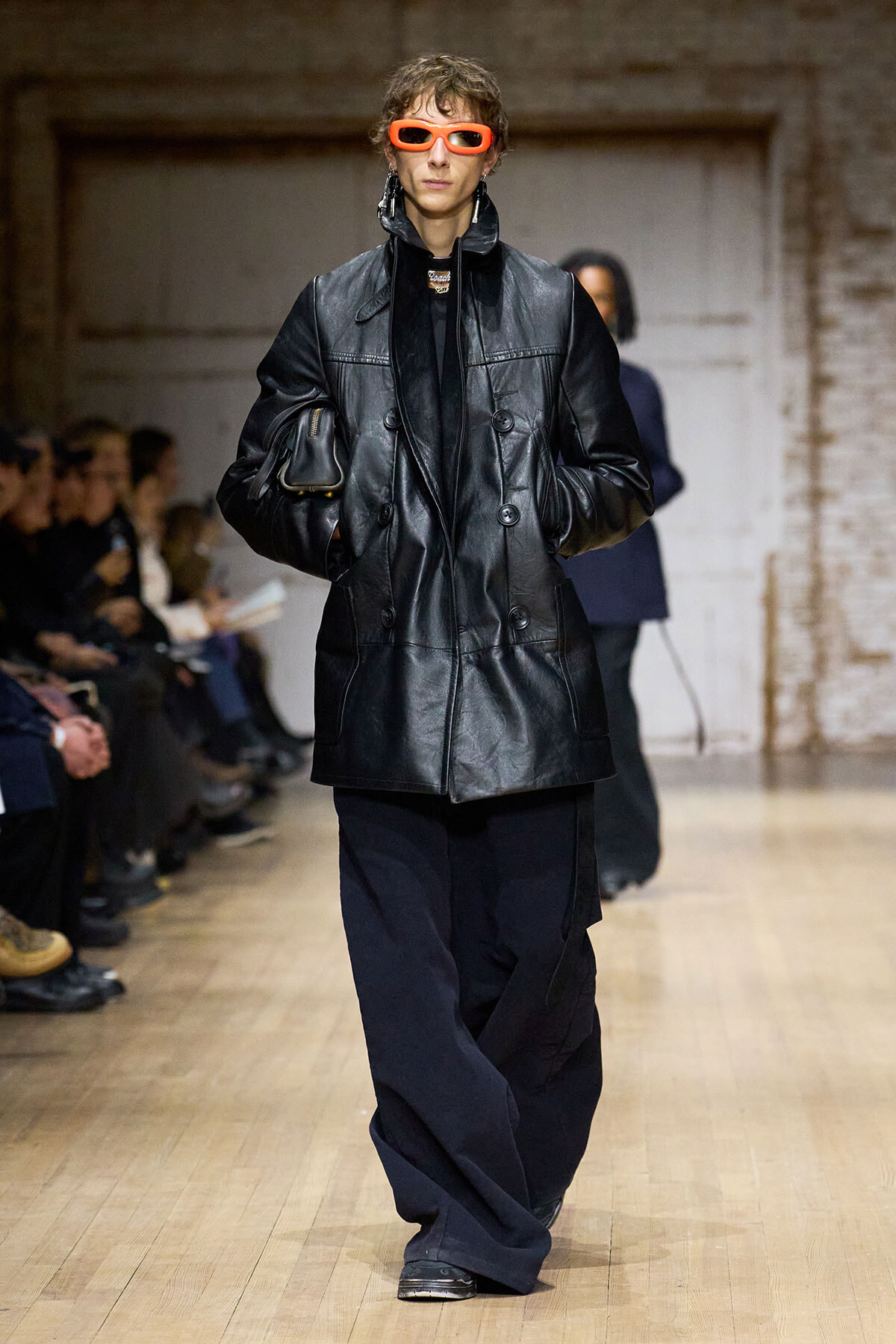 Model walks the runway in a black oversized leather double-breasted coat, bright orange rectangular sunglasses, wide navy pants, and a small bag tucked under the arm.
