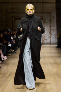 Model walking a fashion runway in a long black coat with fuzzy lining, light-wash jeans, and yellow sunglasses; brick wall backdrop.