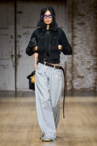 Model walks down runway in a black cropped jacket with fur collar, oversized light-wash jeans, and purple rectangular glasses, holding a small bag.