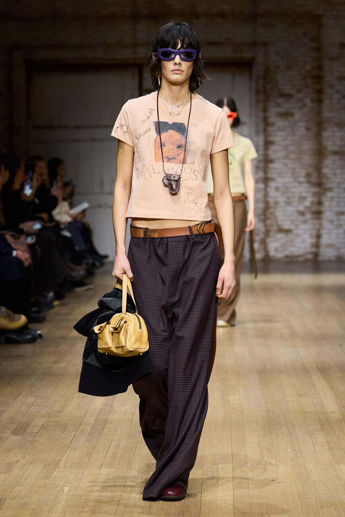 Model on a runways walk in a peach graphic tee, loose plaid trousers, brown belt, purple sunglasses, carrying a tan bag with a coat draped over her arm.