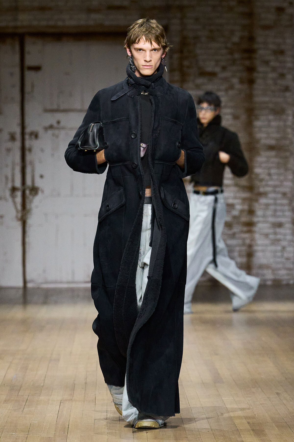 Model walks the runway in a long black coat, holding a small black bag against an industrial brick backdrop.