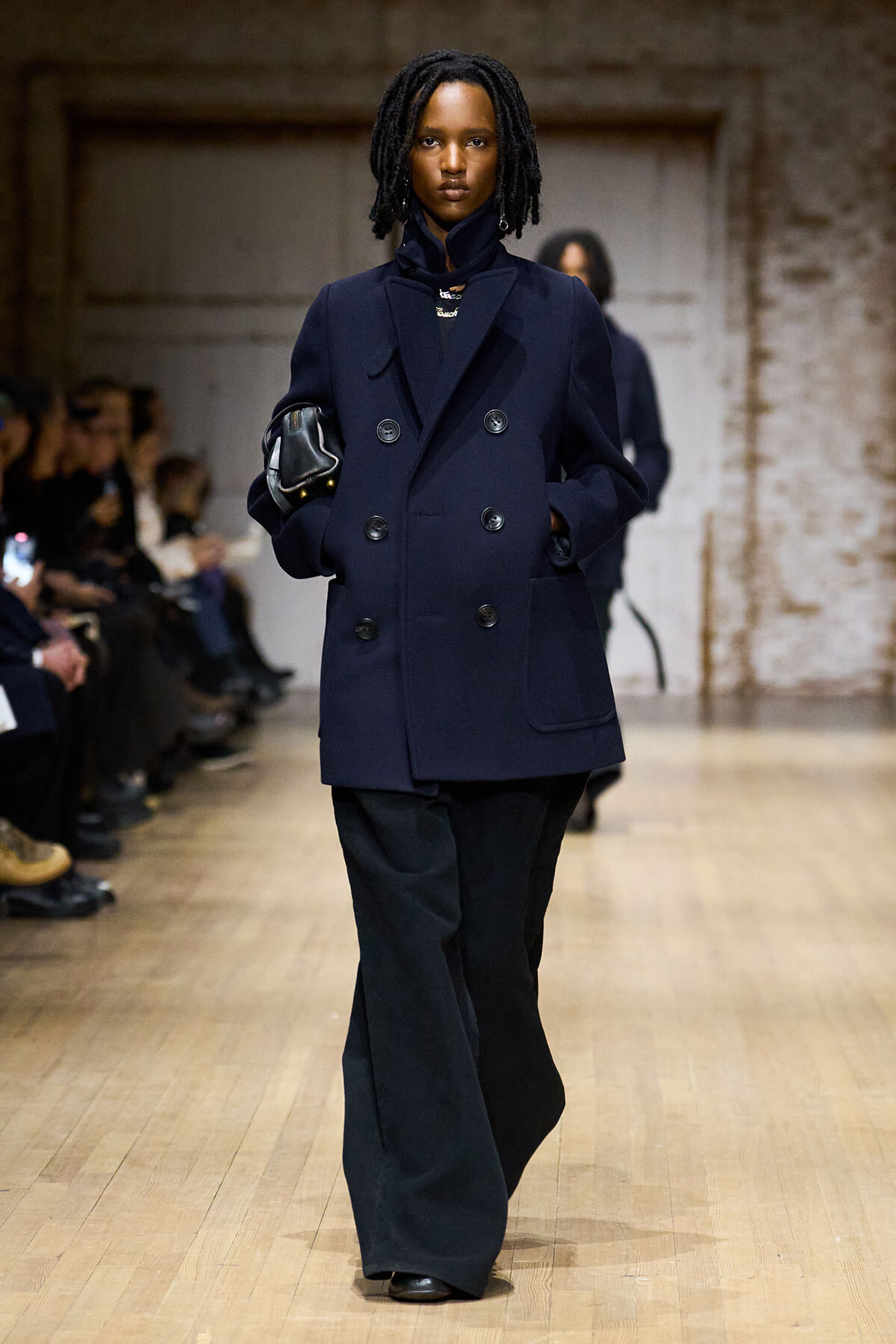 Model on a runway wearing a navy double-breasted coat and wide-leg black pants, carrying a small black bag under her arm.