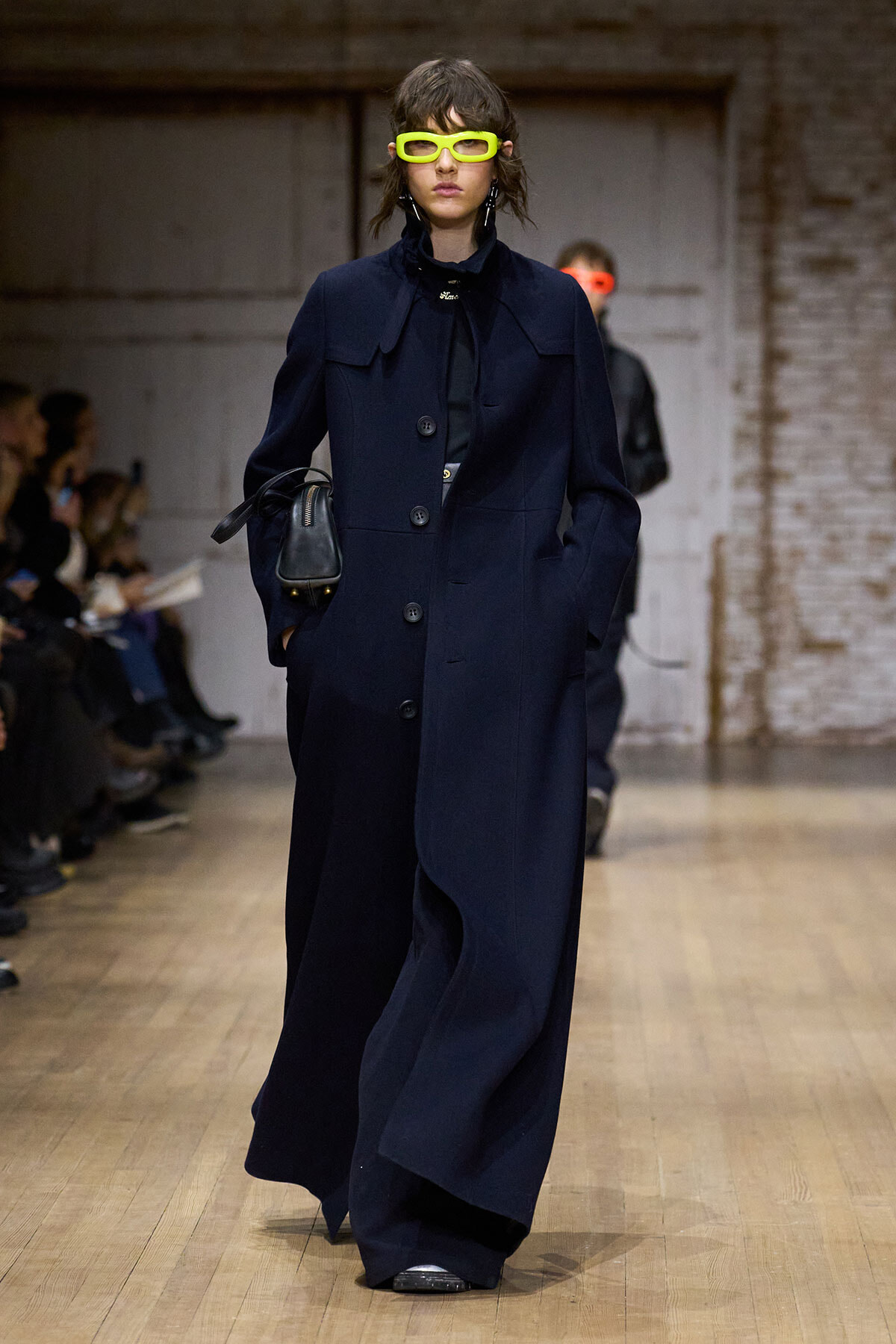 Model on a runway wearing a long navy coat and bright yellow oversized sunglasses, carrying a small black bag; audience visible on the left.