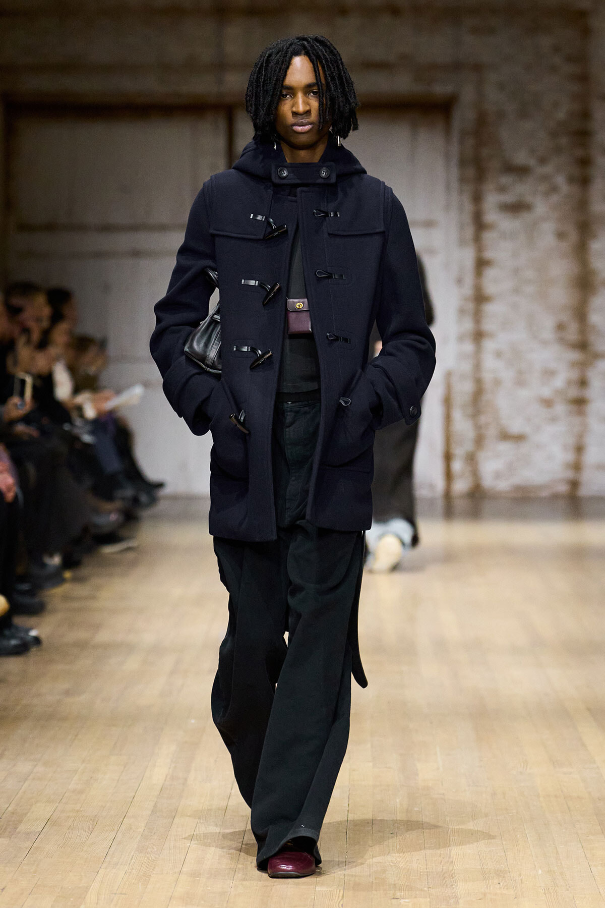 Model walking the runway in a dark duffle coat with toggle closure and wide pockets, audience in background.