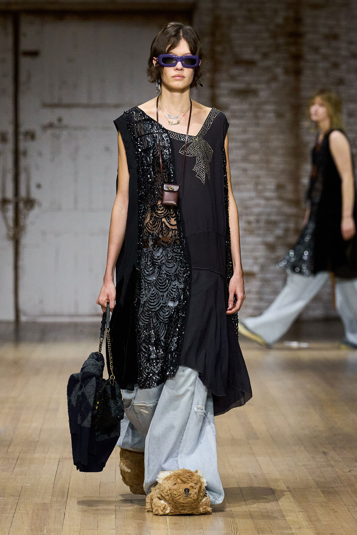 Model walks a runway in a black sequined, asymmetric dress with draped panels, purple sunglasses, and layered necklaces, carrying a small brown bag; a plush teddy bear sits at her feet.